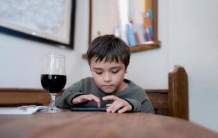 Kid playing game on mobile while waiting for food, Portrait young boy sitting in coffee shop sending text to friends,Child playing game online on cellphone.Children with internet or technology concept photo