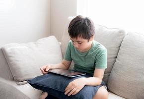 High key portrait young boy playing game on tablet sitting on sofa with light shining from window, Kid playing games on internet, Cinematic portrait Child doing homework online at home photo