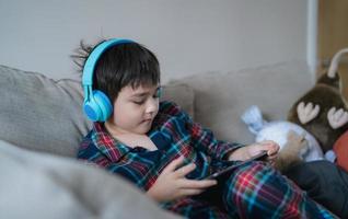 Happy boy wearing headphone listening to music, Kid with smiling face having fun playing game online on tablet with friends on holiday, Child wearing pajamas relaxing at home in the morning photo