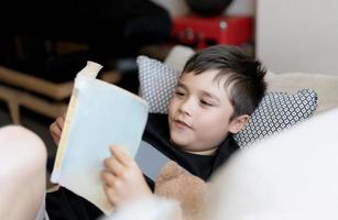 niño de la escuela leyendo un libro para la tarea, niño tiro en la cabeza acostado en el sofá disfruta leyendo la historia en la sala de estar, niño relajándose en casa en la primavera soleada, educación en el hogar, concepto de educación foto