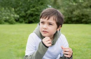 retrato de un niño mirando profundamente en sus pensamientos, un niño de la escuela sentado solo en la hierba verde en el jardín, un niño lindo jugando al aire libre con una naturaleza verde borrosa en el parque foto