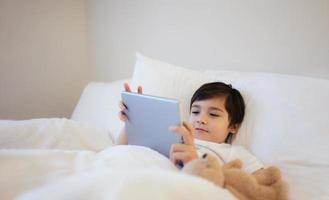 niño feliz acostado en la cama sosteniendo una tableta viendo dibujos animados y charlando con amigos en un bloc digital, un niño lindo jugando juegos en línea en Internet, un niño relajándose por la mañana antes de ir a la escuela foto