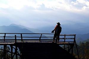 Silhouette of Tourist man standing and holding camera with mountain and blue sky background at view point of Doi Ang Khang, Chiangmai, Thailand. photo