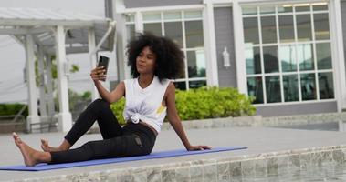 uma mulher vestida para o exercício senta-se em um tapete de ioga falando chamada de vídeo de telefone inteligente. video