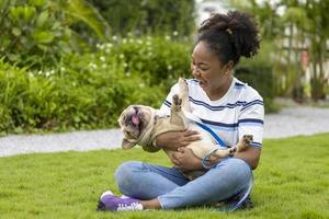 African American woman is playing with her french bulldog puppy while walking in the dog park at grass lawn after having morning exercise during summer photo