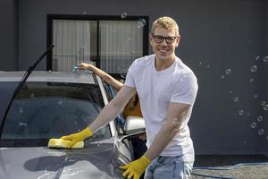 Young Caucasian man is smiling while using sponge and soap water to wash his car in the garage at home photo