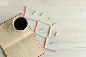 una taza de café, un libro abierto y tizas multicolores sobre un fondo blanco de madera. regreso a la escuela, día mundial del libro. foto