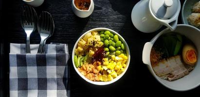 Flat lay of fresh salad with sliced of corn, salmon, green nut or bean,  pineapple, bacon, cucumber and topping with white and black sesame. Set of Healthy food  with spoon and fork put in napkin. photo