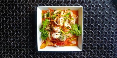 vista superior de la ensalada tailandesa de mariscos picantes con fideos de vidrio, calamares, camarones, rodajas de tomate, cebolla, zanahoria y apio en un plato o plato blanco. endecha plana de comida asiática sobre fondo negro de acero inoxidable. foto