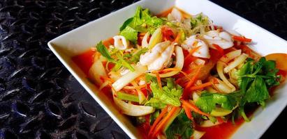 Cierra la ensalada tailandesa de mariscos picantes con fideos de vidrio, calamares, camarones, rodajas de tomate, cebolla, zanahoria y apio en un plato o plato blanco. comida asiática y famosa sobre fondo negro de acero inoxidable. foto