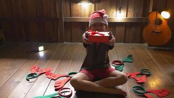 un niño con un sombrero de santa se sienta solo en casa abriendo una caja de regalo el día de navidad.