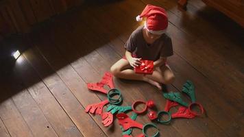 un niño con un sombrero de santa se sienta solo en casa abriendo una caja de regalo el día de navidad. video