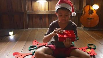 un niño con un sombrero de santa se sienta solo en casa abriendo una caja de regalo el día de navidad.