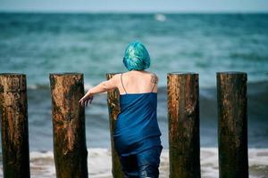 Blue-haired woman performance artist in blue dress standing on beach holding paint brush, back view photo