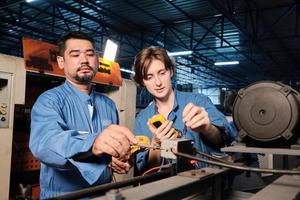 Asian male and White female engineer workers in safety uniforms work by inspecting machines' voltage current, checking, and maintaining at manufacturing factory, electric system service occupations. photo