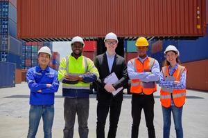 Group of multiracial workers team in safety uniforms, arms crossed and looking at camera at logistics terminal with many stacks of containers, loading shipping goods, cargo transportation industry. photo