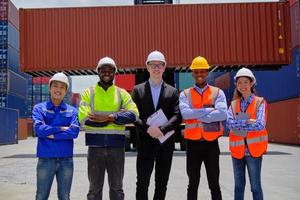 Group of multiracial workers team in safety uniforms, arms crossed and looking at camera at logistics terminal with many stacks of containers, loading shipping goods, cargo transportation industry. photo