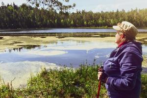 Side view aged senior woman stand hold red nordic sticks and enjoy panoramic view of lake in sunny beautiful day. Texture and nature well-being concept background photo