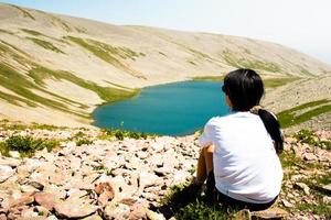 Back view young caucasian woman alone sit enjoy blue Levani lake panorama in tranquil mountains on hike rocky outdoors. Georgia caucasus travel adventure concept