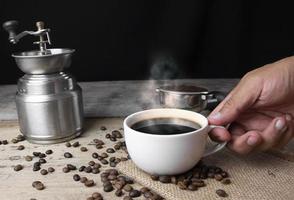 vista frontal mano sosteniendo una taza de café americano negro sobre una mesa de madera con un montón de granos de café arábica asados sobre fondo negro. foto