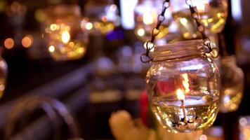 Burning small white wax candles in high transparent glass hanging with trembling flame on blurry background in ball room close up view. video