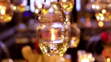 Burning small white wax candles in high transparent glass hanging with trembling flame on blurry background in ball room close up view. video