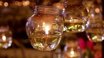 Burning small white wax candles in high transparent glass hanging with trembling flame on blurry background in ball room close up view. video