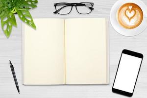 un cuaderno en blanco abierto, un teléfono inteligente con bolígrafo y una taza de café con leche en una mesa de madera. foto