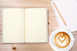 An open notebook with pencil and a cup of coffee on wooden table. Latte art coffee on top photo