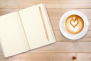 An open notebook with pencil and a cup of coffee on wooden table. Latte art coffee on top photo