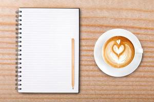 An open blank notebook with pencil and a cup of coffee on wooden table. Latte art coffee on top photo