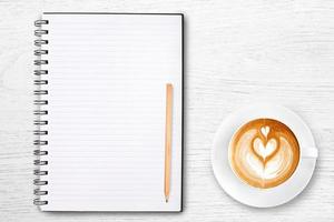 An open blank notebook with pencil and a cup of coffee on wooden table. Latte art coffee on top photo