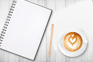 An open blank notebook with pencil and a cup of coffee on wooden table. Latte art coffee on top photo