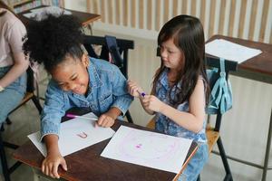 Preschool Girl Kid Drawing With Color Pencil On White Paper On Table In Classroom With Friends photo