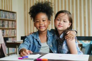 Preschool Girl Kid Drawing With Color Pencil On White Paper On Table In Classroom With Friends photo