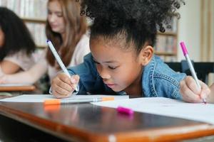 Preschool Girl Kid Drawing With Color Pencil On White Paper On Table In Classroom With Friends photo