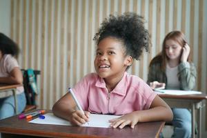 Preschool Girl Kid Drawing With Color Pencil On White Paper On Table In Classroom With Friends photo