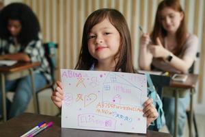 niña preescolar dibujando con lápiz de color sobre papel blanco en la mesa en el aula con amigos foto