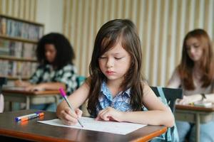 Preschool Girl Kid Drawing With Color Pencil On White Paper On Table In Classroom With Friends photo