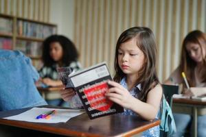 Preschool Girl Kid Drawing With Color Pencil On White Paper On Table In Classroom With Friends photo
