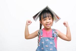 Back to school. Smiling little girl carrying a backpack holding books on her head looking at the camera on a white background with copy space. Girl glad ready to study. photo