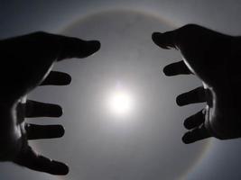 Beautiful photograph of the sun with a circular rainbow surrounded by a bright sky and white clouds with shadows of hands reaching out. Phenomenon, sun halo. photo