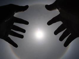Beautiful photograph of the sun with a circular rainbow surrounded by a bright sky and white clouds with shadows of hands reaching out. Phenomenon, sun halo. photo