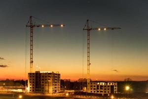 grúas torre de silueta y edificios altos de varios pisos sin terminar en construcción al atardecer en el desierto en un sitio de construcción iluminado foto