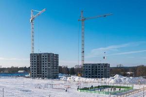 Tower cranes and unfinished multi-storey high near buildings under construction site in sunny winter day with a view of the children's sports and hockey grounds photo