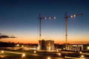 grúas de torre y altura de varios pisos inacabada cerca de edificios en construcción en la noche de la puesta de sol con un espectacular fondo de nubes de colores foto