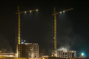grúas torre y edificios altos de varios pisos sin terminar en construcción por la noche en un sitio de construcción iluminado foto