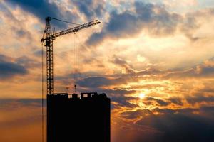 silueta de grúas torre y edificios altos de varios pisos inacabados en construcción en la puesta de sol por la noche en los rayos del sol poniente foto