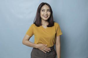 Pleased cheerful Asian woman keeps hand on belly feels full after delicious dinner dressed casually stands thoughtful against blue background. photo