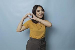 Attractive young Asian woman feels happy and romantic shapes heart gesture expresses tender feelings wears casual yellow t-shirt against blue background. People affection and care concept photo